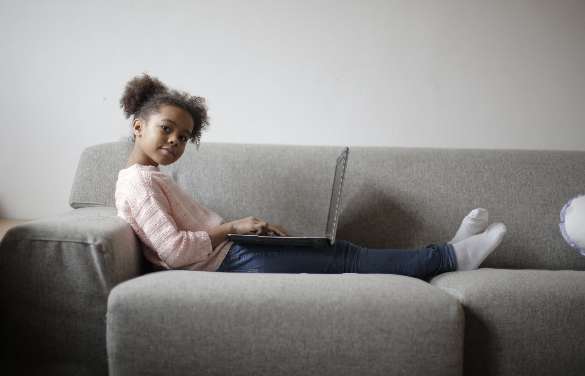 Petite fille aux cheveux frisés assise sur un canapé avec un ordinateur portable sur les genoux pour illustrer la facilité de partager des choses en ligne sur Internet