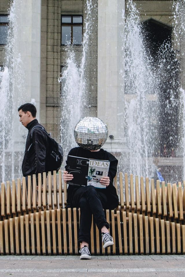 Homme assis sur un banc publique tenant un journal à la main dont le titre est "Ideas" (idées en Français). L'homme a la tête en forme de boule à facette.